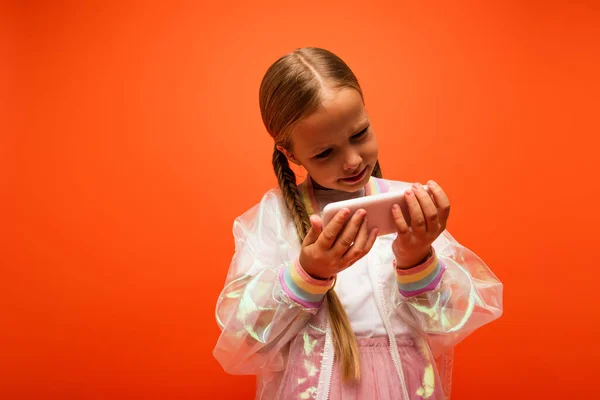 Mädchen in durchsichtiger Regenjacke blickt auf Handy isoliert auf Orange — Stockfoto