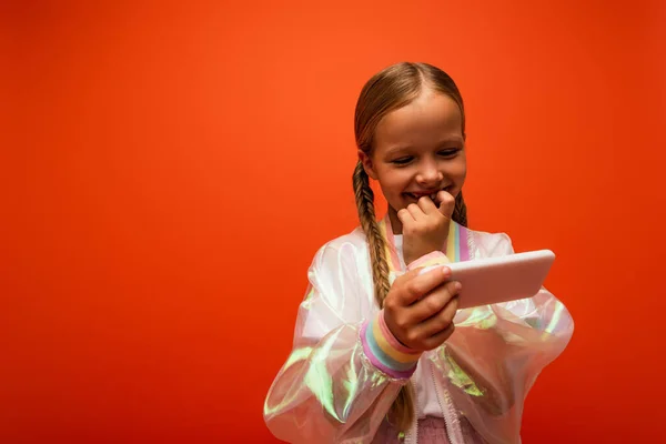 Chica sonriente en chaqueta de lluvia cogida de la mano cerca de la boca mientras mira el teléfono inteligente aislado en naranja - foto de stock