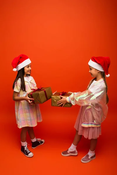 Повна довжина усміхнених дев'ятнадцяти дівчат у капелюхах Санти, що тримають різдвяні подарунки на помаранчевому фоні — стокове фото