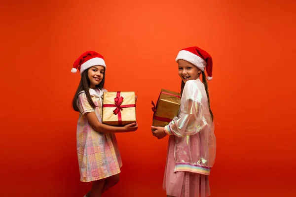 Cheerful girls in santa hats holding gift boxes and smiling at camera isolated on orange — Stock Photo