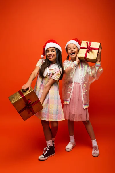 Pleine longueur de filles à la mode en chapeaux santa posant avec des cadeaux de Noël sur fond orange — Photo de stock