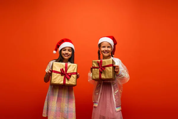 Amigos felices en sombreros de santa sonriendo a la cámara mientras sostiene cajas de regalo aisladas en naranja - foto de stock