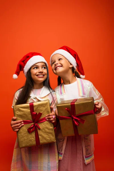 Pleased kids in santa hats holding christmas presents isolated on orange — Stock Photo