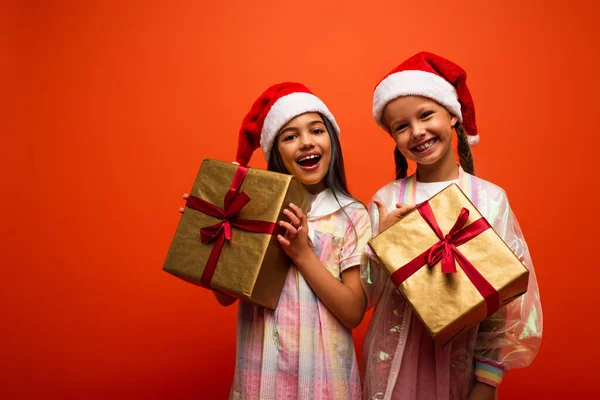 Amazed and happy girls in santa hats standing with gift boxes and looking at camera isolated on orange — Stock Photo