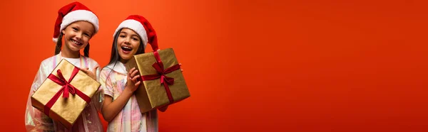 Cheerful preteen girls in santa hats holding christmas gifts isolated on orange, banner — Stock Photo