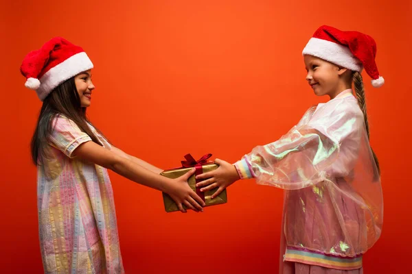 Side view of smiling girl in santa hat presenting gift to happy friend isolated on orange — Stock Photo