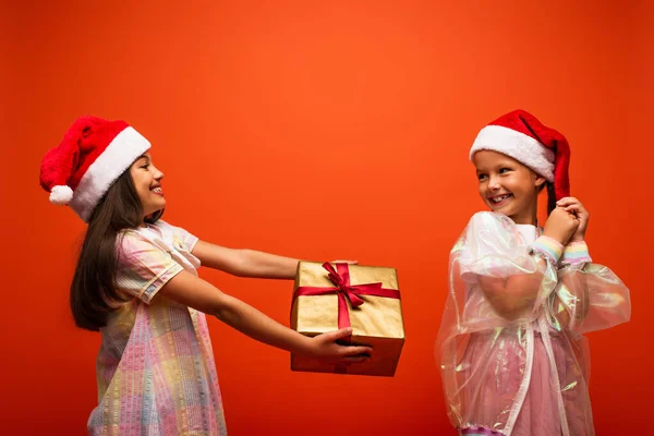 Smiling girl in santa hat presenting christmas gift to pleased friend isolated on orange — Stock Photo