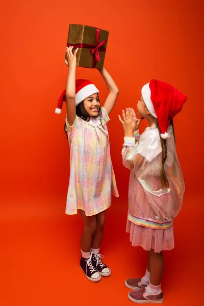 Piena lunghezza di allegra ragazza in cappello di Babbo Natale tenendo regalo di Natale vicino amico battendo le mani su sfondo arancione — Foto stock