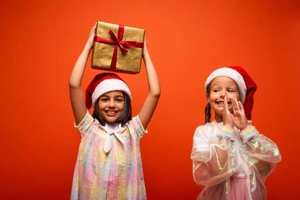 Aufgeregtes Kind mit Weihnachtsmannmütze klatscht Hände neben Mädchen mit Geschenkbox in erhobenen Händen isoliert auf orange — Stockfoto