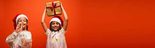 Heureuse fille dans santa chapeau tenant boîte cadeau dans les mains levées près ami excité applaudissant mains isolées sur orange, bannière — Photo de stock