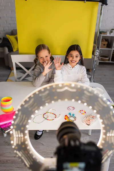 Blogueiros alegres acenando mãos perto de contas na mesa e câmera digital com anel de luz em primeiro plano borrado — Fotografia de Stock