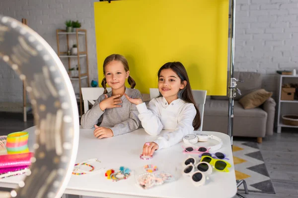 Blogueurs vidéo souriants parlant près de différents accessoires sur la table devant la lampe circulaire floue — Photo de stock