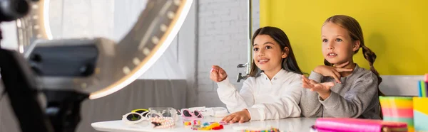 Cheerful bloggers talking and gesturing near different accessories on table and blurred ring light, banner — Stock Photo