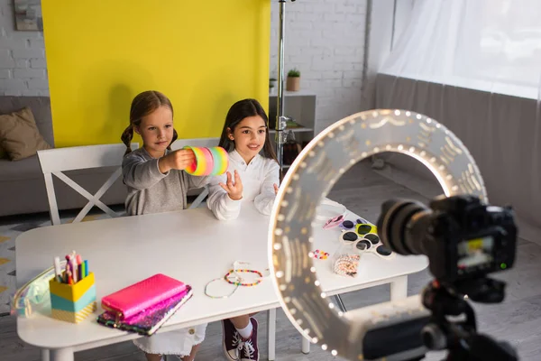 Sonriente video blogger hablando cerca de un amigo demostrando un juguete en espiral frente a una lámpara de círculo borroso con cámara digital - foto de stock