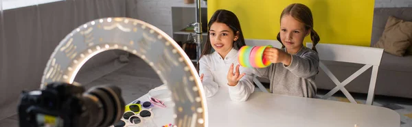 Preteen girl showing spiral toy near blogger talking in front of blurred digital camera in circle lamp, banner — Stock Photo