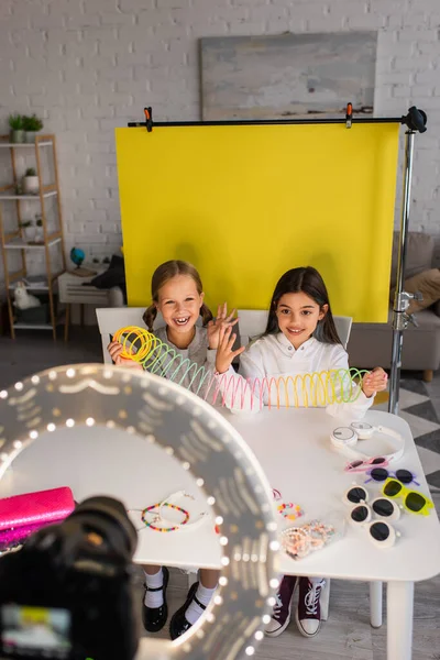 Blogueurs joyeux parler et montrer jouet en spirale sur fond jaune en face de l'appareil photo numérique floue — Photo de stock