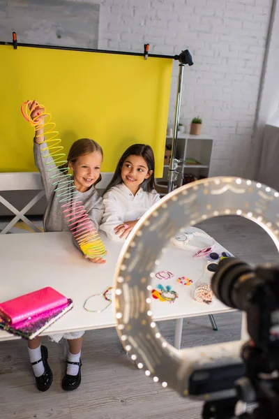 Menina sorrindo mostrando brinquedo espiral enquanto sentado perto de amigo e câmera digital borrada em lâmpada de círculo — Fotografia de Stock