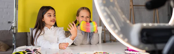Blogueiro morena falando perto de menina sorridente mostrando brinquedo espiral na frente da lâmpada círculo borrada, banner — Fotografia de Stock