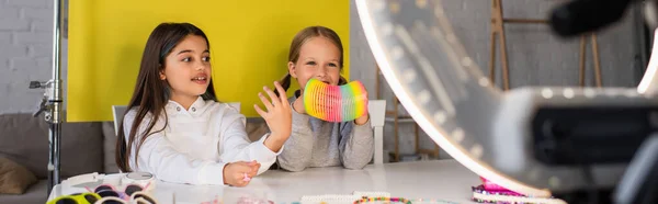 Brunette blogger talking near smiling girl showing spiral toy in front of blurred circle lamp, banner — Stock Photo