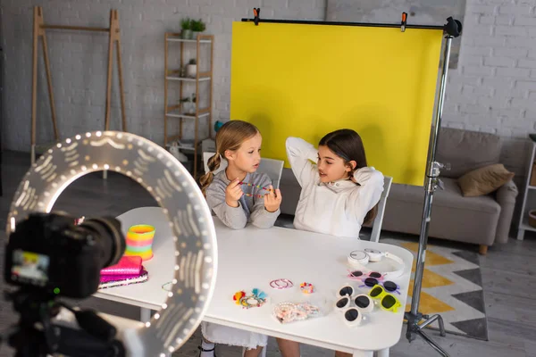 Girl showing beads near video blogger on yellow background in front of blurred digital camera with circle lamp — Stock Photo