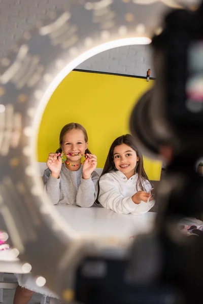 Smiling video blogger showing beads near friend and digital camera in circle lamp on blurred foreground — Stock Photo