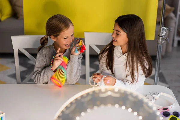 Blogueiros de vídeo alegres demonstrando contas e brinquedo em espiral perto da lâmpada do círculo em primeiro plano desfocado — Fotografia de Stock