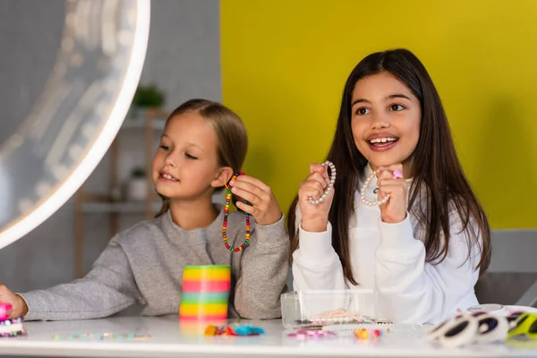 Blogueurs souriants montrant des perles et des bracelets près de la lampe circulaire sur le premier plan flou — Photo de stock