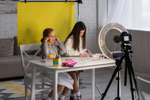 Preteen video blogger talking near girl and different accessories on table in front of digital camera in ring light — Stock Photo