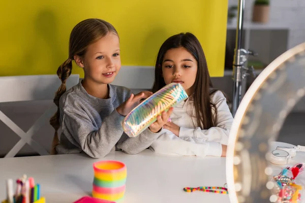 Preteen blogger demonstrating pencil case near friend and circle lamp on blurred foreground — Stock Photo