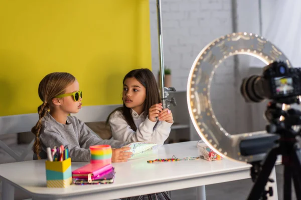 Blogueiro de vídeo em óculos de sol segurando caixa de lápis e falando com amigo perto de câmera digital borrada em lâmpada de círculo — Fotografia de Stock