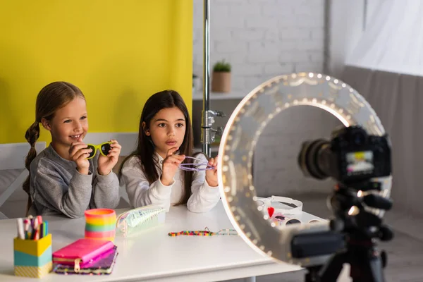 Preteen bloggers showing stylish sunglasses on yellow background in front of blurred digital camera — Stock Photo