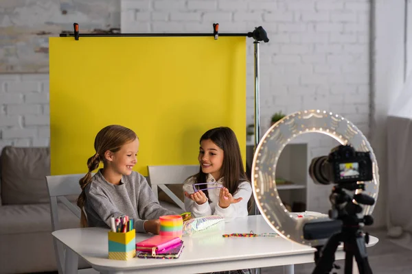 Meninas pré-adolescentes alegres mostrando óculos de sol durante a gravação de vídeo blog na câmera digital borrada perto de fundo amarelo — Fotografia de Stock