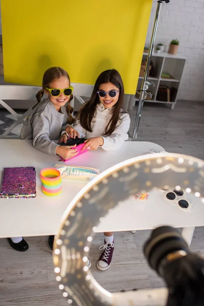 Alegres video bloggers en gafas de sol de moda mostrando estuche de lápiz cerca de la cámara digital borrosa sobre fondo amarillo en casa - foto de stock