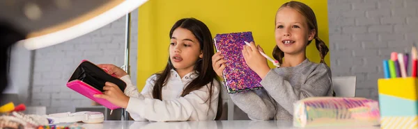 Bloggers preadolescentes demostrando diferentes artículos de papelería cerca de la lámpara de círculo en primer plano borroso, pancarta - foto de stock