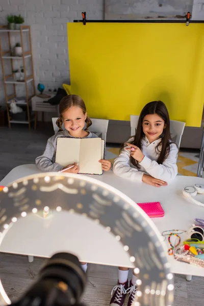 Blogger vidéo souriant pointant vers un ami montrant un carnet vierge devant une lampe circulaire floue avec appareil photo numérique — Photo de stock