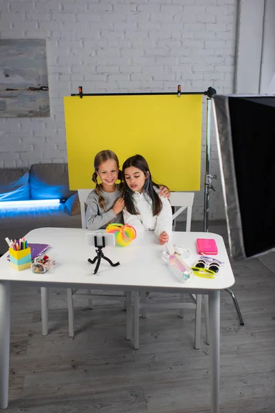 Happy girl embracing friend near sunglasses and stationery on table while recording video blog at home — Stock Photo