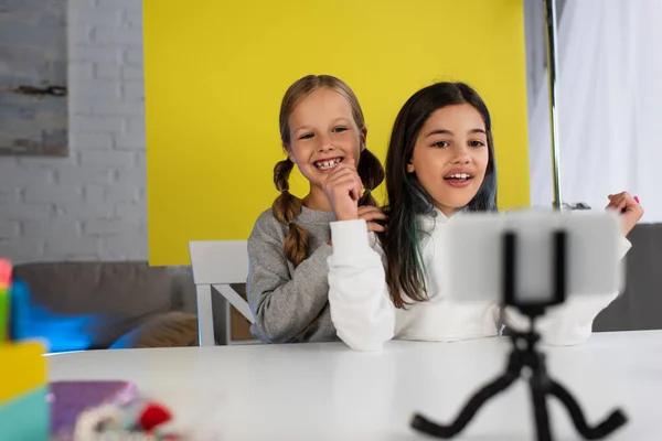 Blogueurs vidéo joyeux parler tout en regardant le téléphone mobile et assis sur fond jaune à la maison — Photo de stock