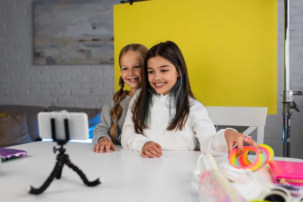 Heureux blogueurs préadolescents regardant smartphone flou sur le titulaire tout en étant assis sur fond jaune à la maison — Photo de stock