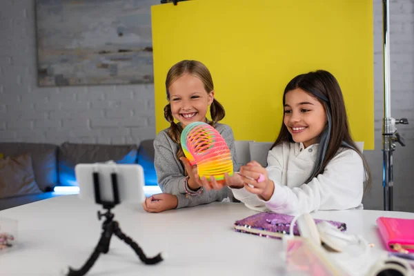 Cheerful video bloggers showing spiral toy on yellow background in front of mobile phone at home — Stock Photo