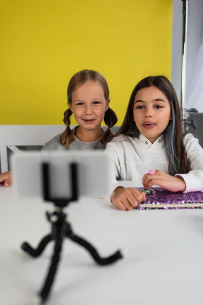 Happy preteen bloggers sitting on yellow background and looking at blurred mobile phone on holder — Stock Photo