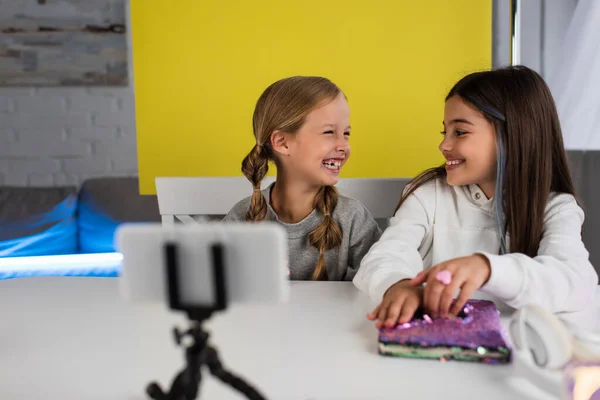 Cheerful bloggers looking at each other near blurred mobile phone on holder at home — Stock Photo