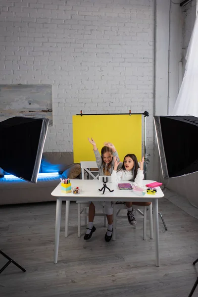 Excited girls gesturing while recording video blog on mobile phone near lamps and yellow background at home — Stock Photo