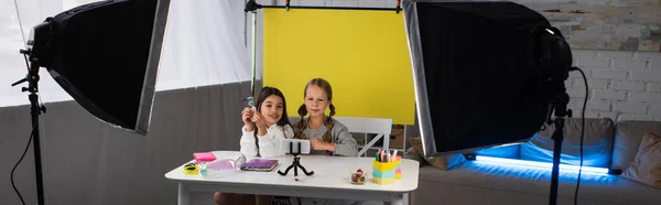 Preteen friends recording video blog on cellphone on yellow background near lamps at home, banner — Stock Photo