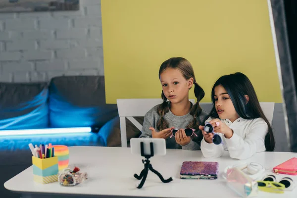 Preteen bloggers demonstrating trendy sunglasses near copybook and various accessories in front of mobile phone on holder — Stock Photo