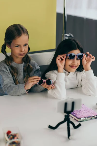 Preteen bloggers demostrando gafas de sol con estilo cerca de smartphone en titular - foto de stock