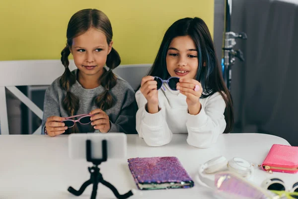 Blogueurs préadolescents montrant des lunettes de soleil à la mode près de carnet brillant et écouteurs devant le téléphone mobile sur le titulaire — Photo de stock