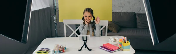 Vídeo blogger acenando mão perto de telefone celular e acessórios diferentes no fundo amarelo em casa, banner — Fotografia de Stock