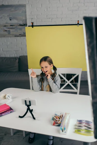 Blogueiro feliz apontando com o dedo perto do telefone móvel no suporte e fundo amarelo em casa — Fotografia de Stock