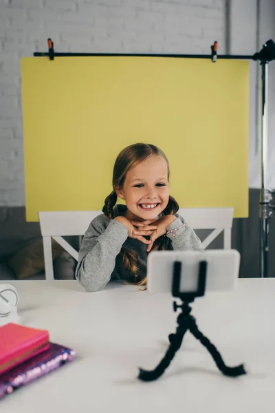 Glückliche jugendliche Bloggerin lächelt über verschwommenes Handy auf Halterung auf gelbem Hintergrund zu Hause — Stockfoto