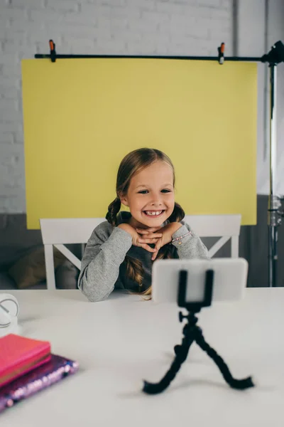 Happy preteen blogger smiling at blurred mobile phone on holder on yellow background at home — Stock Photo